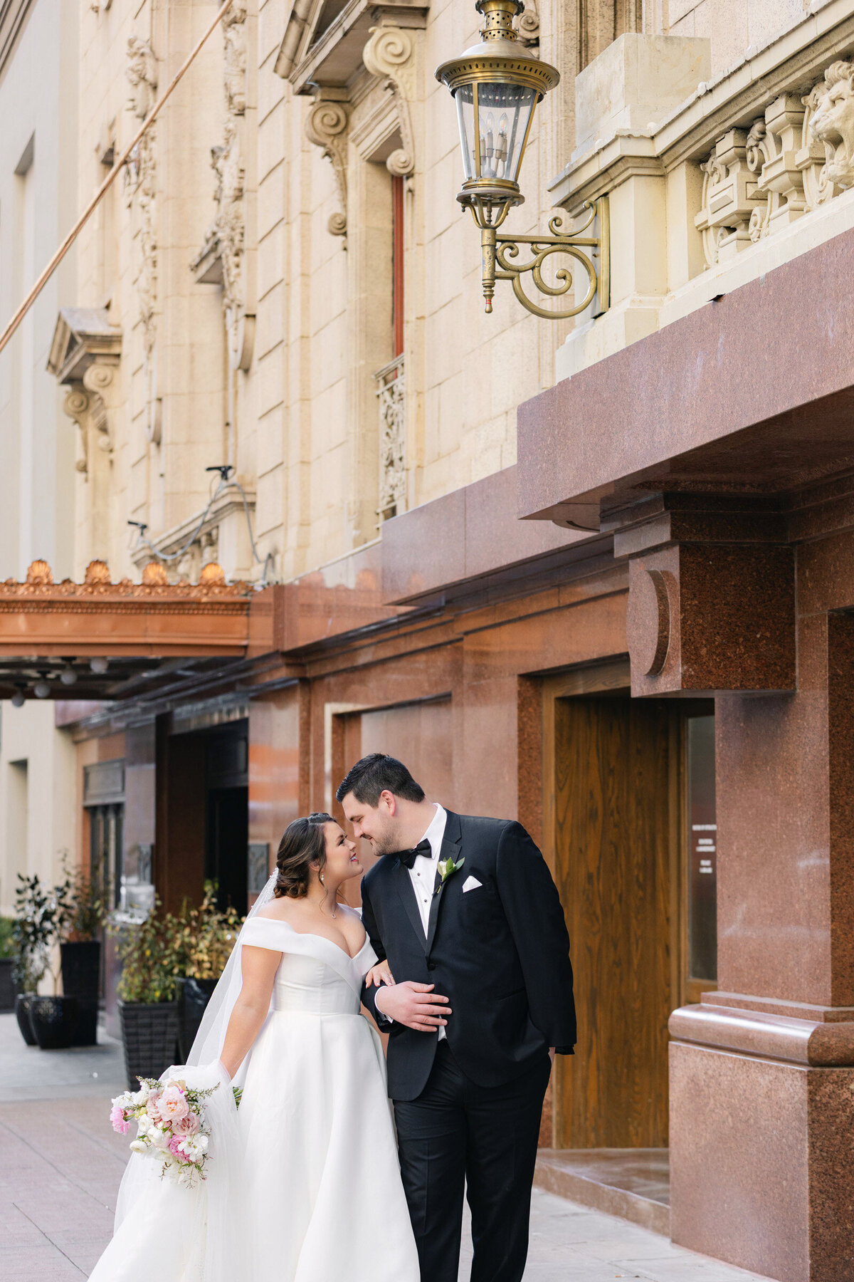 bride and groom snuggling outside The Adolphus in Dallas with the hotel’s architecture behind them, capturing an intimate and romantic wedding moment.