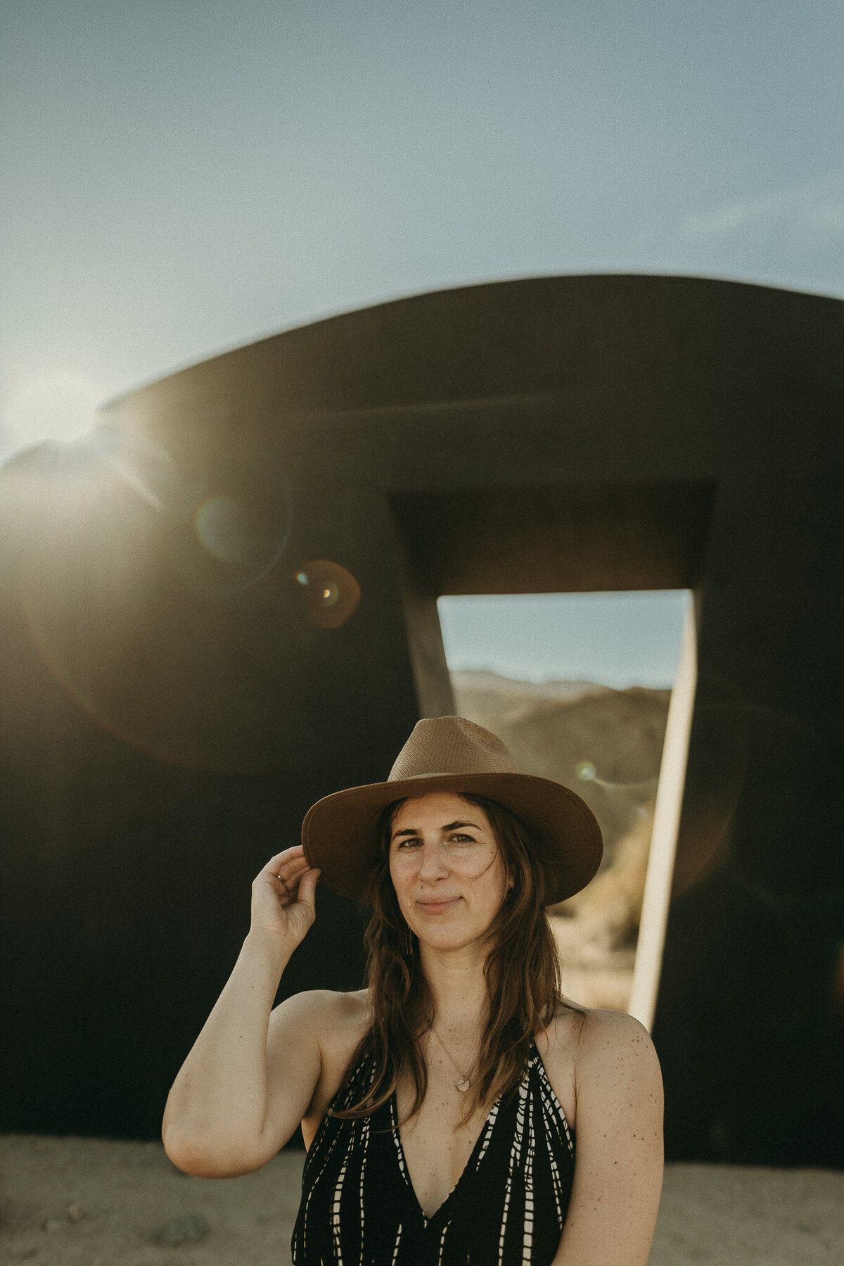 Creative headshot of bay area photographer holding sun hat in front of desertx installation