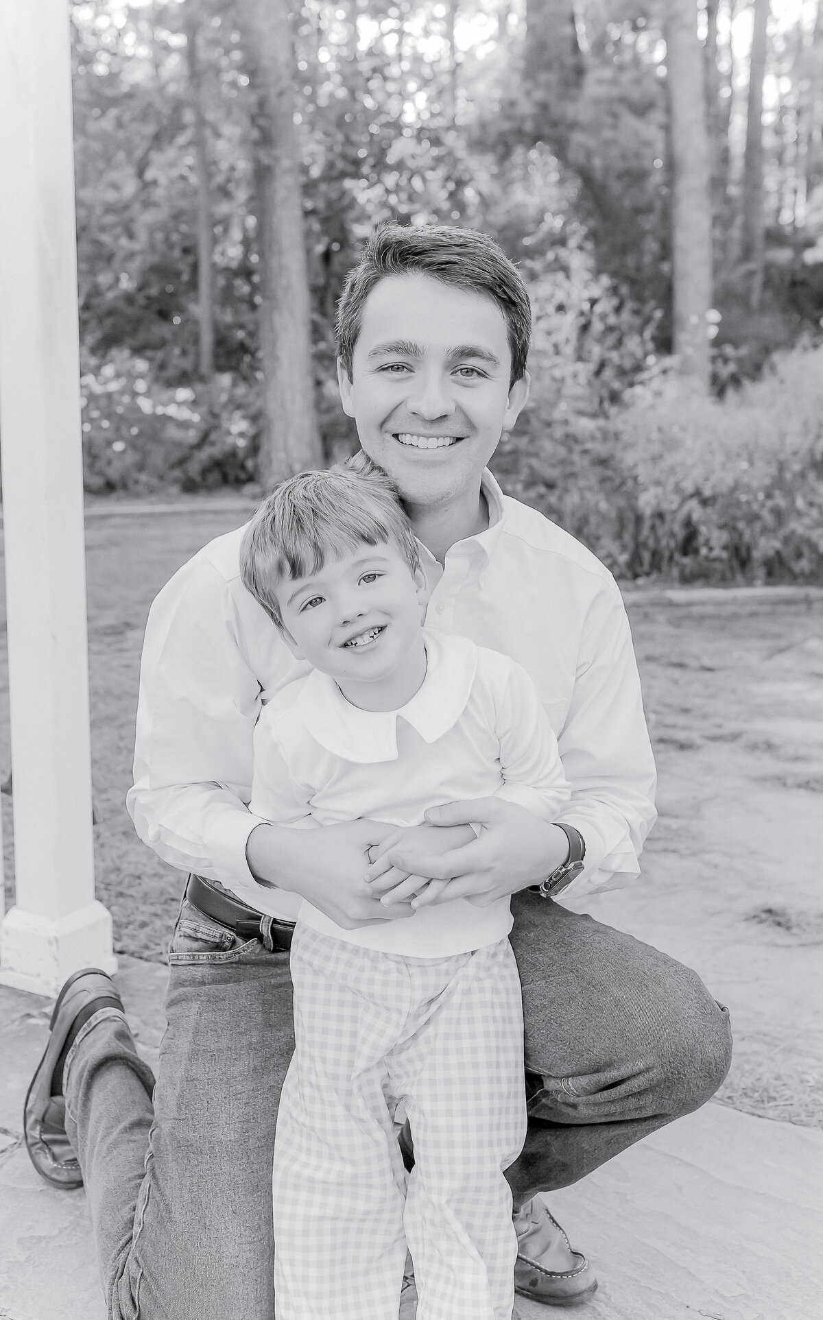 Black and white portrait of a father kneeling and embracing his young son, both smiling warmly. Their close bond and joyful expressions capture the love and connection of family, reflecting the timeless and heartfelt style of a Birmingham family photographer