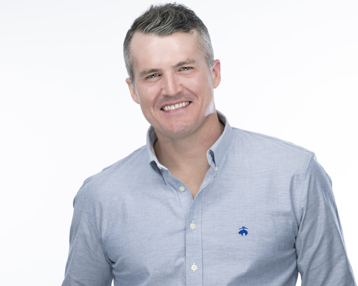 Bright, modern headshot in MA of a man wearing a light blue button-down shirt, smiling warmly against a white background, by Rob Faber Photography, specializing in headshots in MA and CT.