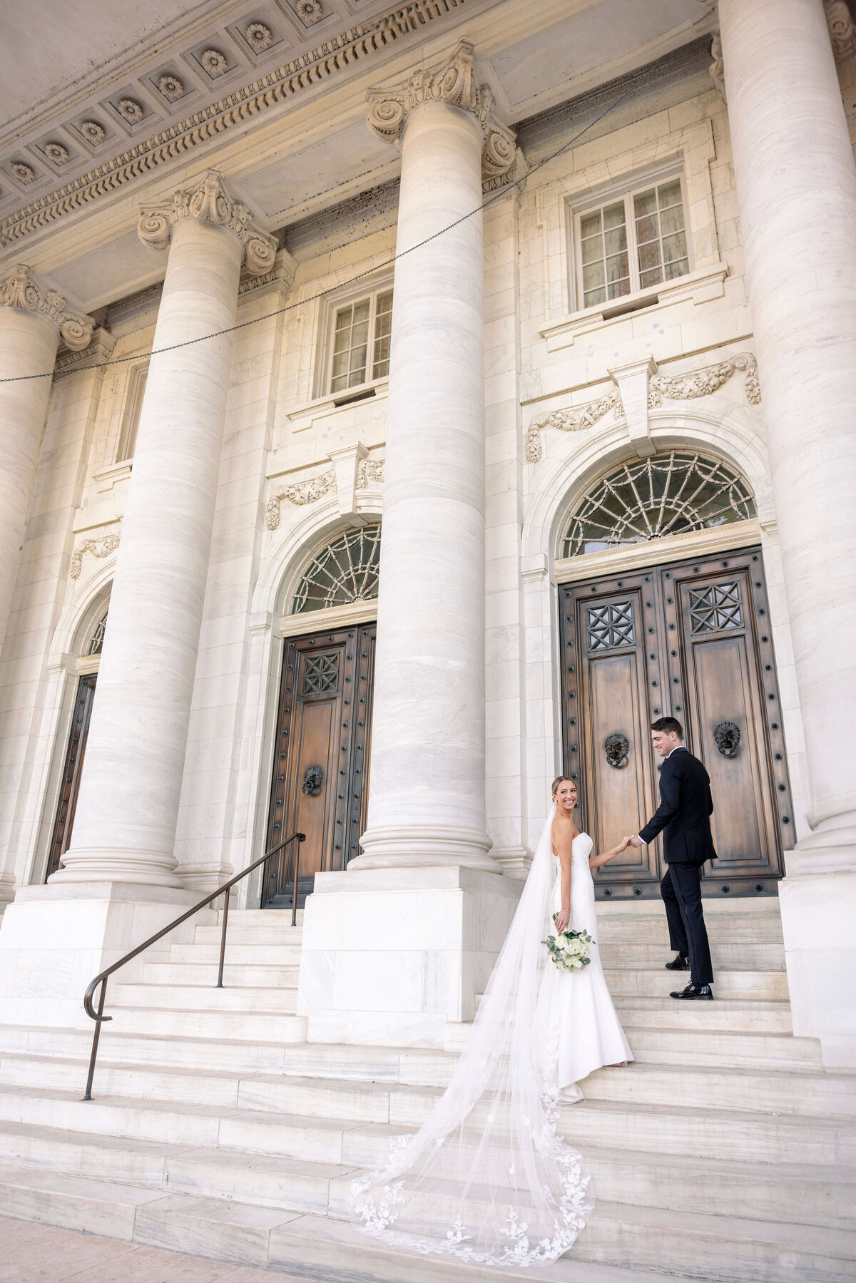 A bride in a white gown with a long, lace-trimmed veil stands with a groom in a black suit on the steps of a grand building with large wooden doors and tall columns. The groom looks back, holding her hand.