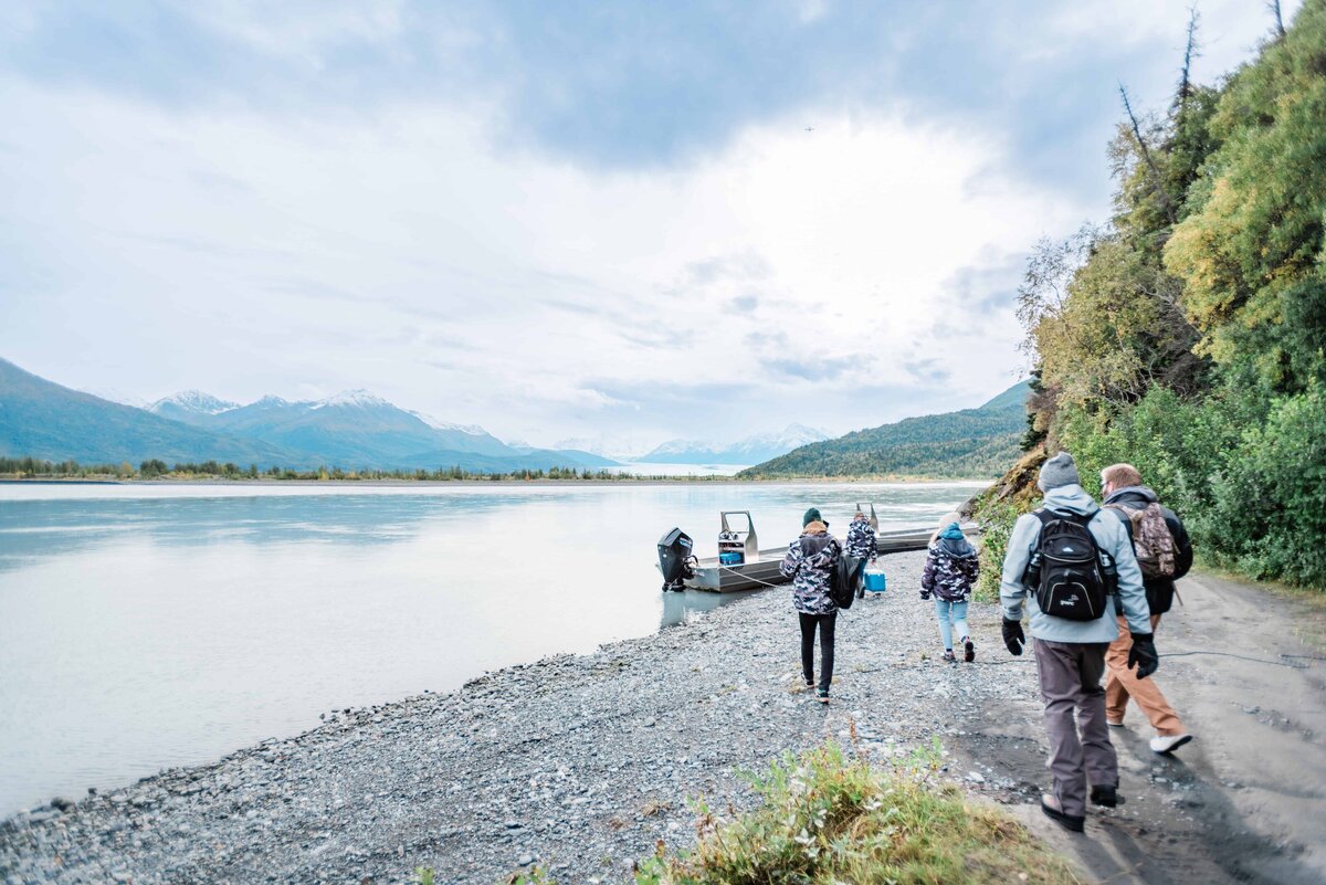 A family heads out to a flat bottom boat ride for a nature tour during a 2 day adventure luxe glacier elopement package in Talkeetna, Alaska.  They are dressed in warm coats and hats, and mountains are snow covered, rising in the distance beyond the river's edge. 