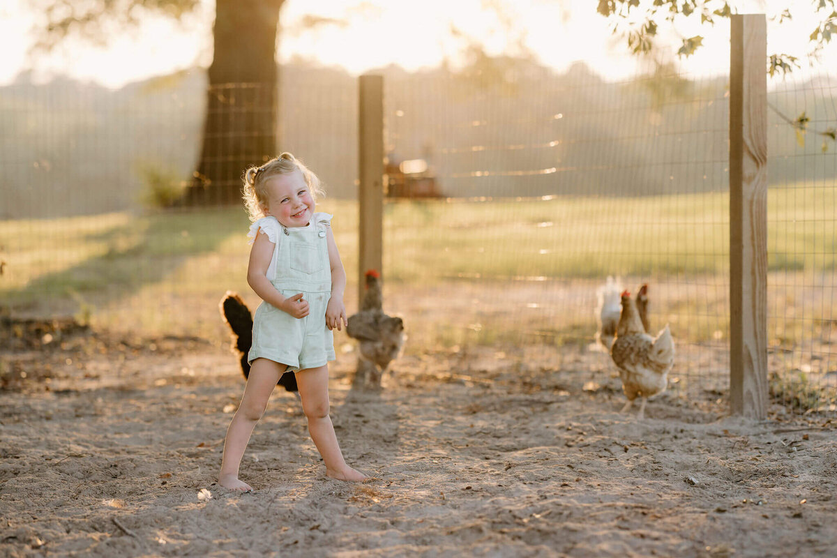candid photo of 3 year old girl in white shirt and green overalls standing with chickens at East Texas family farm