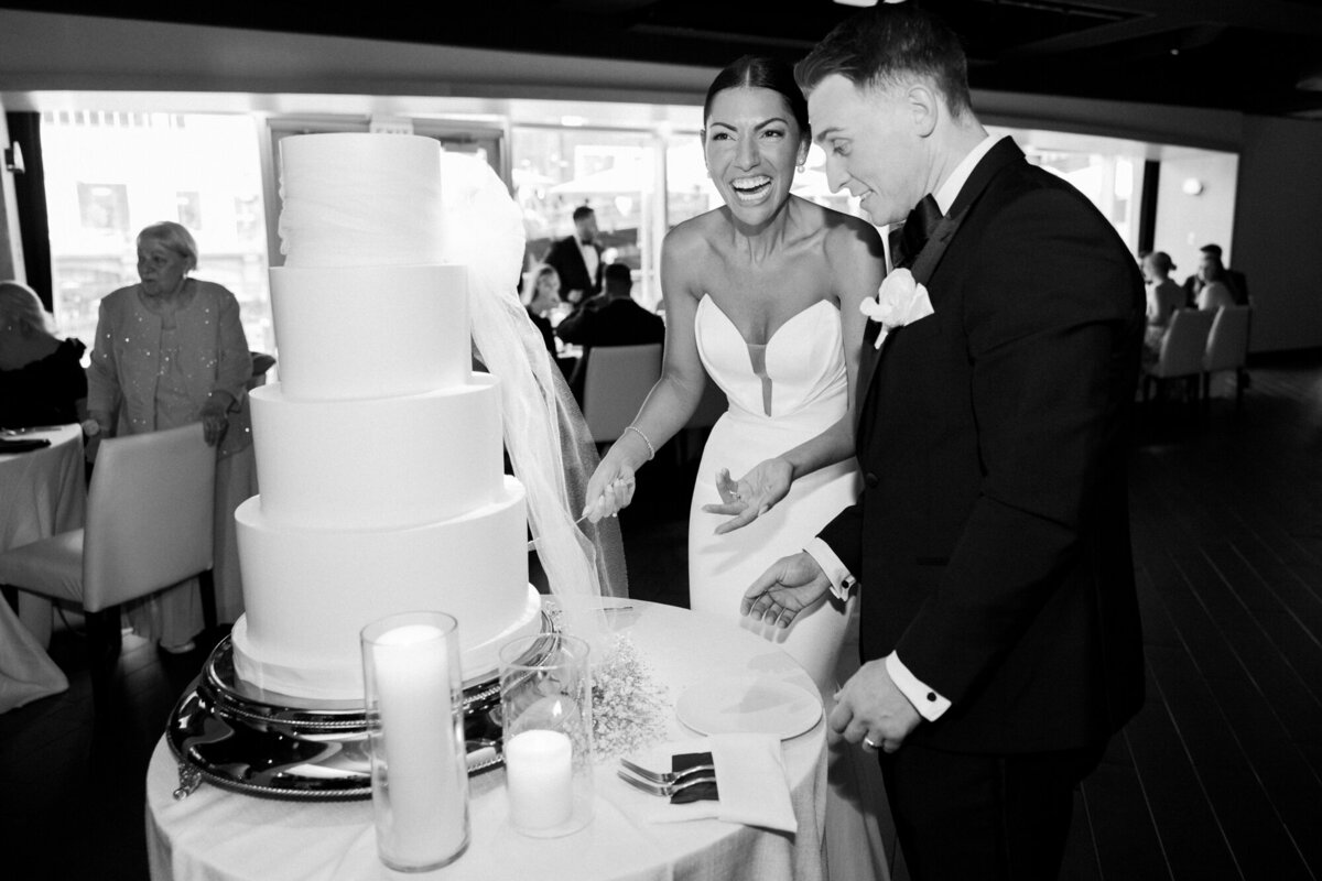 A joyful black and white wedding photo of a couple laughing together as they cut their wedding cake, capturing a lighthearted reception moment.