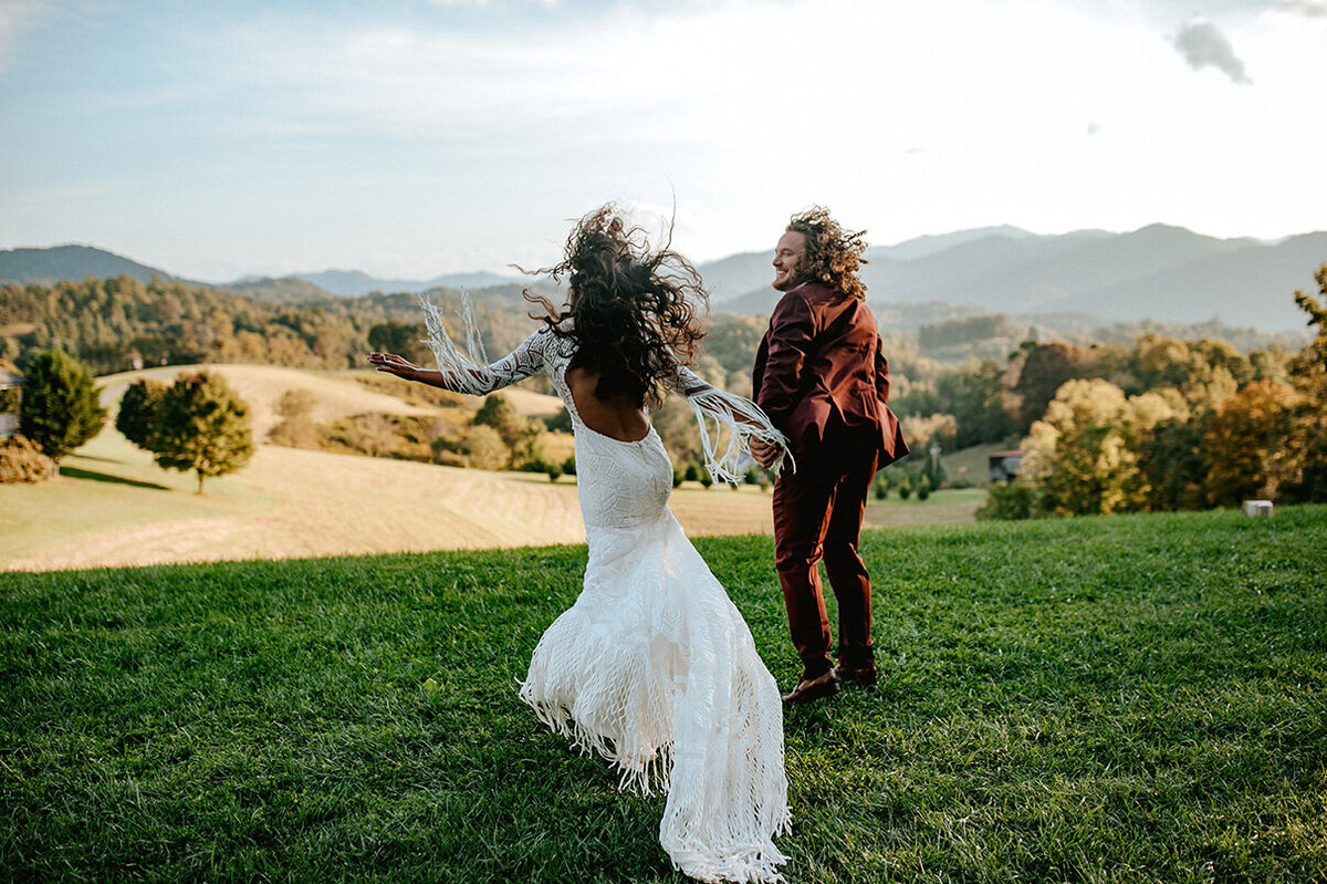 bride and groom wearing a white gown  and burgundy tuxedo running in a mountain field