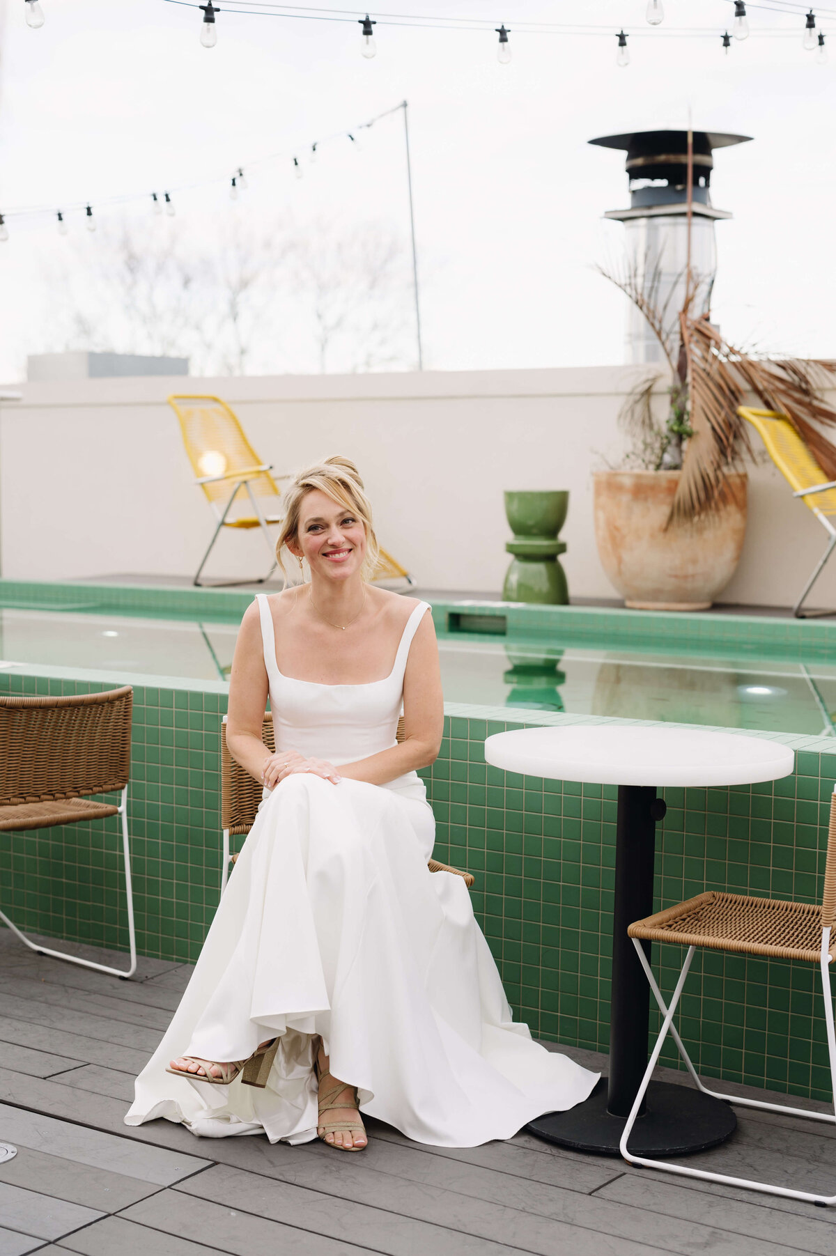 bride in a satin wedding dress sits on a chair next to a green tile pool at common house richmond wedding venues with her hands on her lap smiling