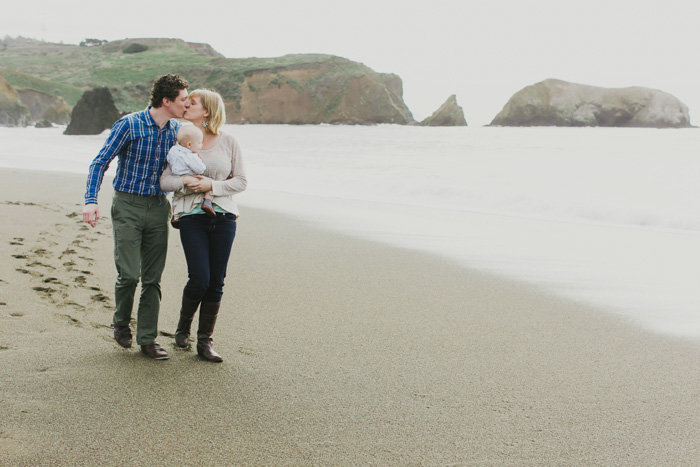 Wide angle shot of mom and dad holding child sharing a kiss as they walk on the beach
