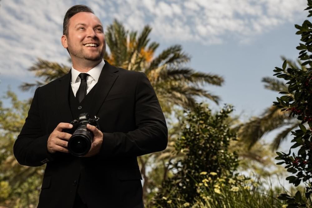 Man-in-a-suit-holding-a-camera-smiling-under-a-sunny-sky-with-palm-trees-in-the-background