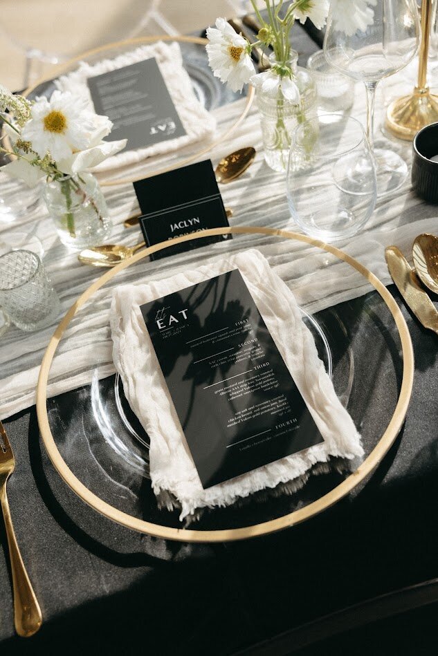 A close-up of an elegant guest table at a National Arts Centre wedding, featuring black linens, a cream cheesecloth runner, gold-rimmed acrylic charger plates, and stylish black menus layered on top. The table is adorned with gold cutlery, bud vases of white flowers, and sophisticated black placecards.