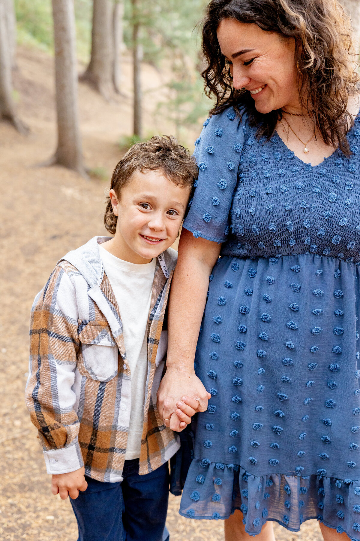 A young boy in a plaid flannel jacket holds his mother’s hand as they walk along a dirt path in the woods. His mother, wearing a blue textured dress, smiles down at him lovingly.