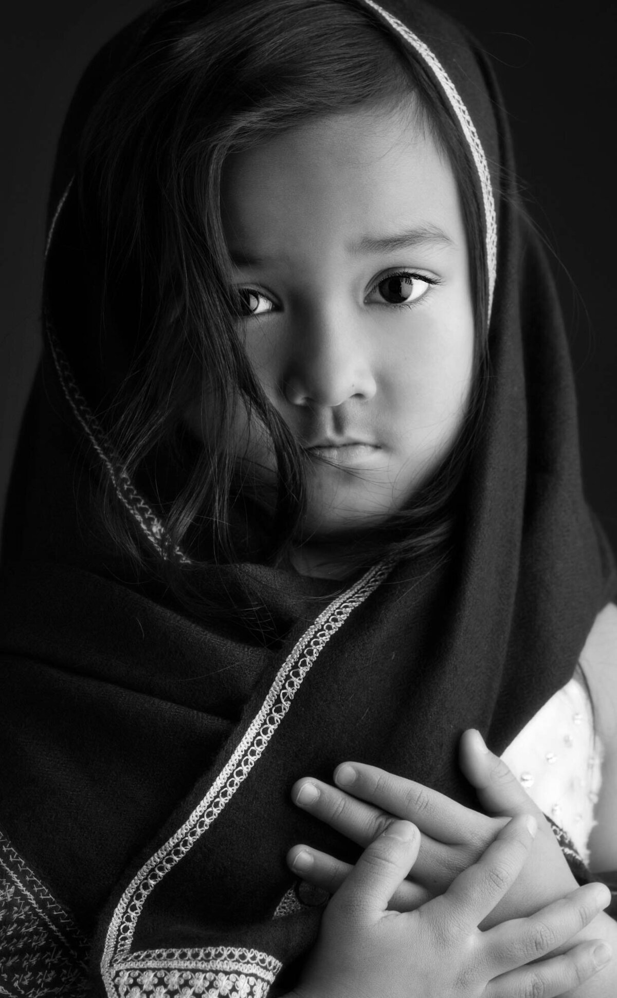 A black and white portrait of a young child with long hair, wrapped in a patterned shawl. The expression is somber, with hands clasped near the chest.