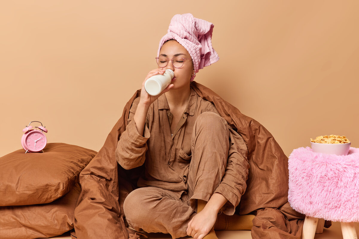 indoor-shot-young-woman-dressed-comfortable-pajama-towel-wrapped-head-drinks-fresh-milk-pose-blanket-poses-bed-near-pillows-has-quick-breakfast-isolated-beige-background
