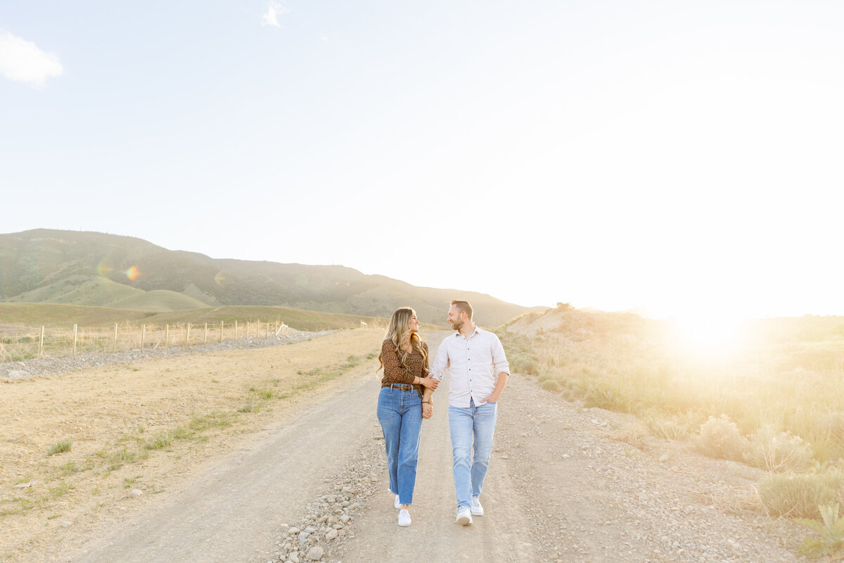 A couple walks hand in hand down a dirt path in Saratoga Springs, Utah, as the sun sets behind the rolling hills. Their love and connection are evident in their gaze toward each other