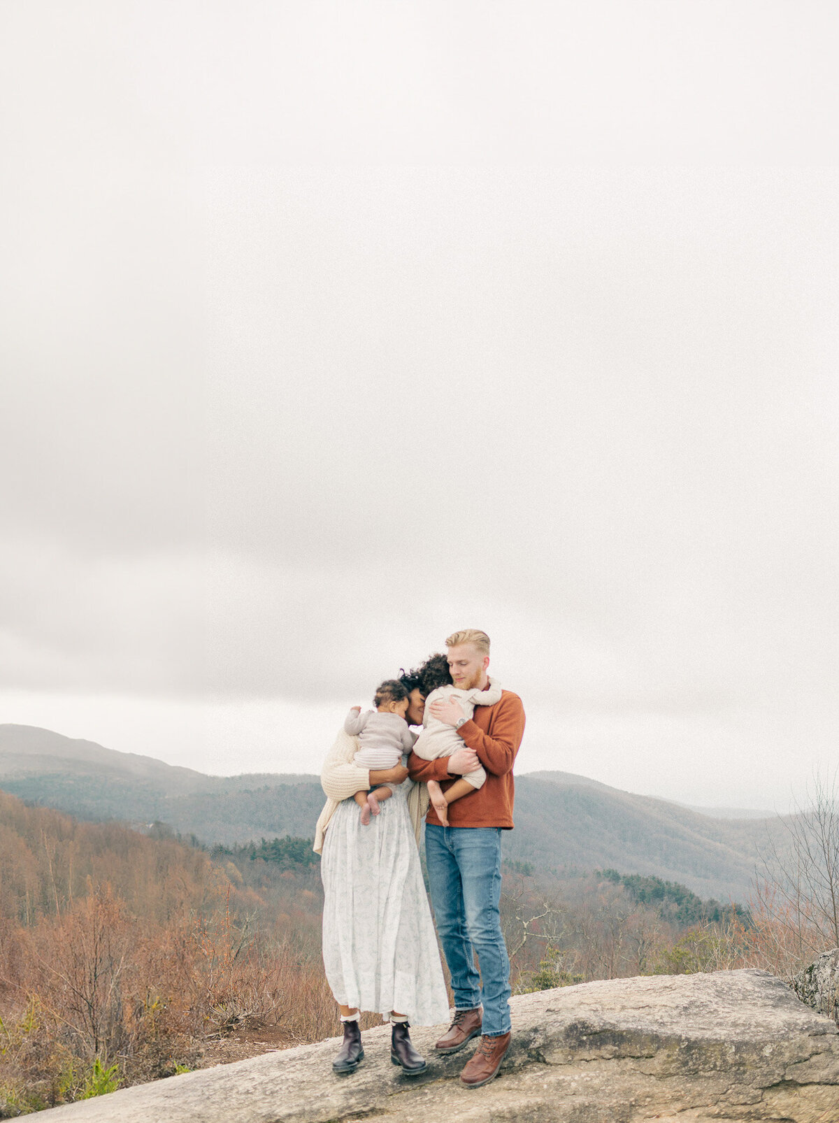 Family of four stands together in neutral outfits during an outdoor newborn session at a Kansas City scenic overlook.
