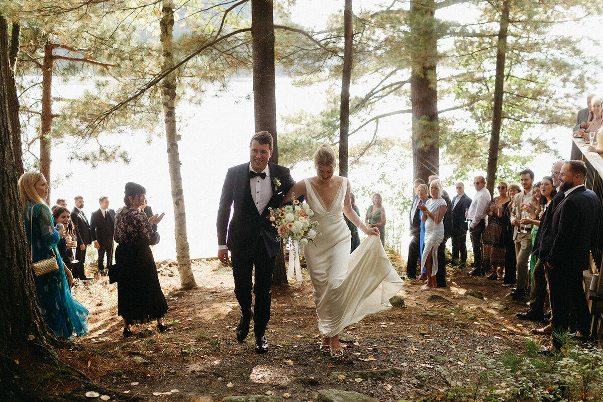 A smiling couple in wedding attire strolls through a sunlit wooded area, their guests applauding as they pass. A serene lake peeks through the trees, all captured beautifully by a Toronto wedding photographer.