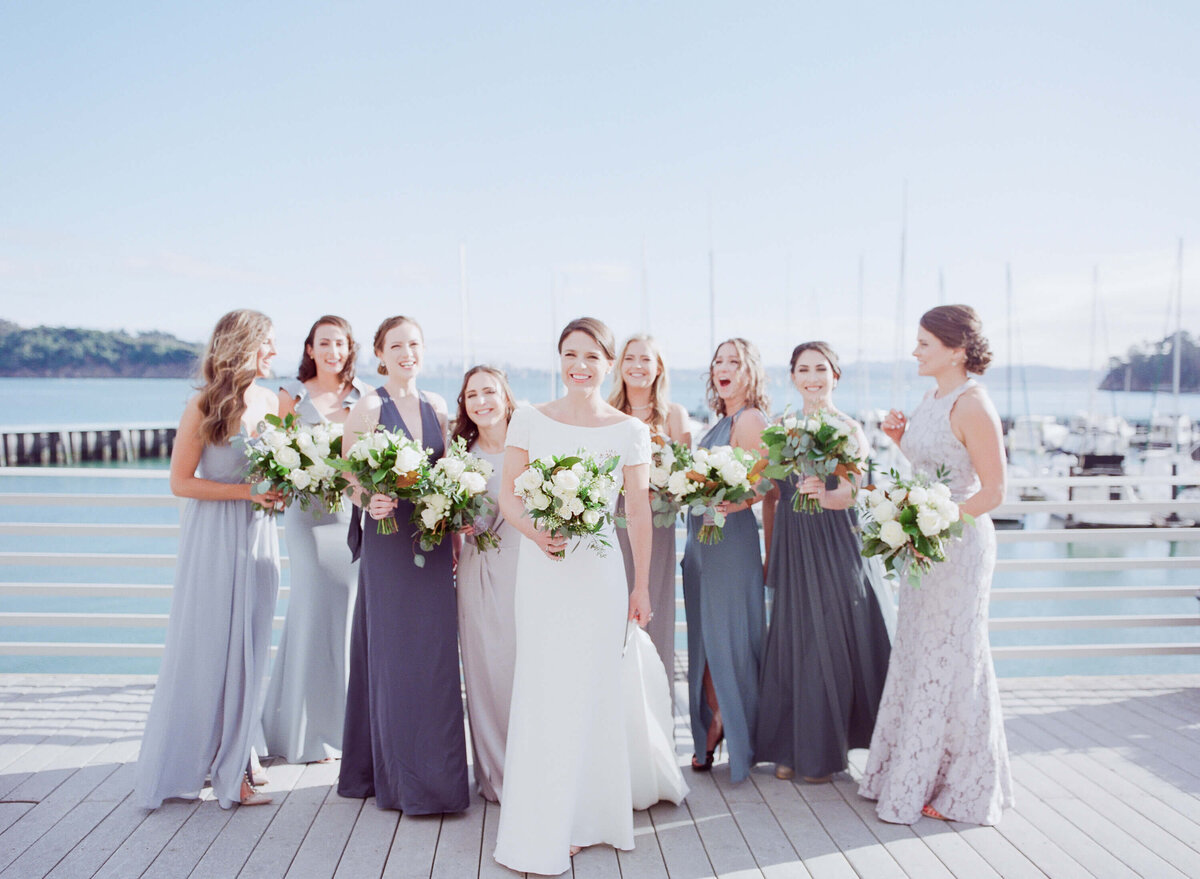 Bride and her bridesmaids hold white flower bouquets pose at an ocean bay.