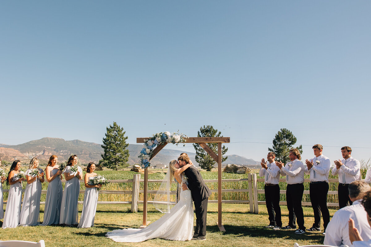 First kiss at an outdoor ceremony, with the wedding party and mountains behind