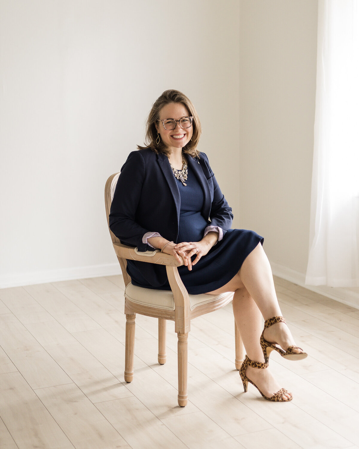 Brunette blonde pregnant female sitting in a chair with legs crossed, wearing glasses, a dark blue dress with a long sleeve dark blue jacket, brown and black cheetah style print heels, smiling at the camera