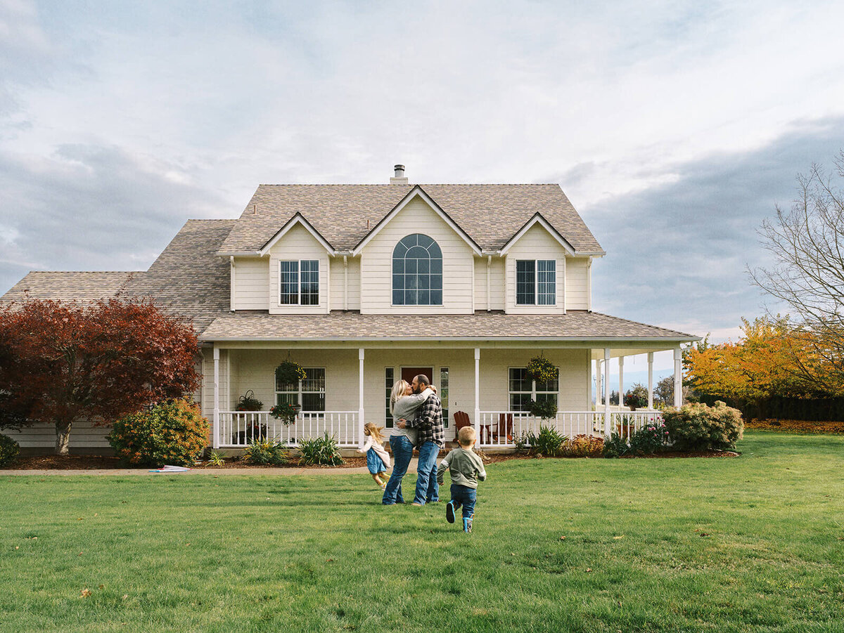 A couple kisses in front of their house while two kids play around them.