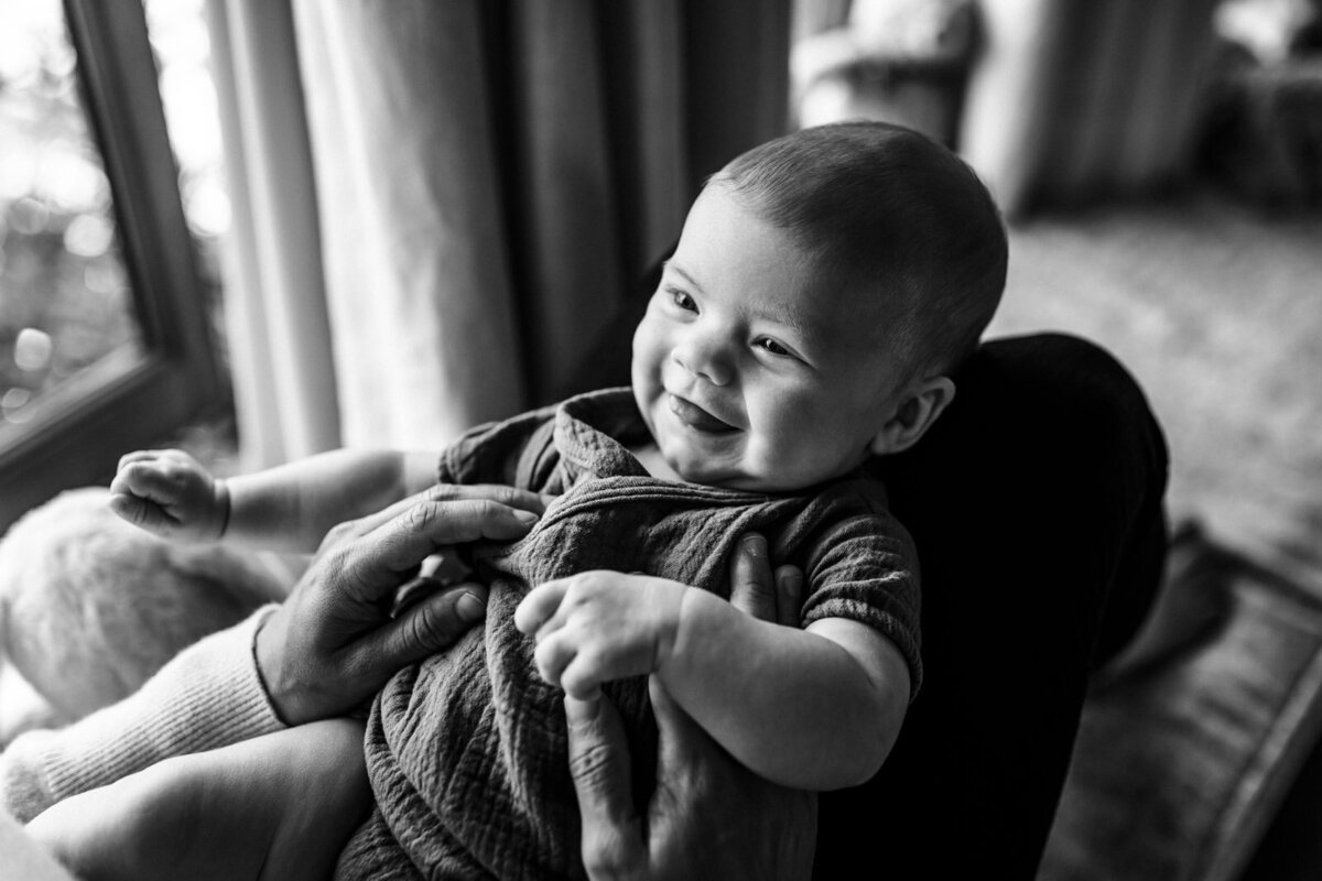 baby sitting on mom's legs on window bench seat and looking and smiling at mom
