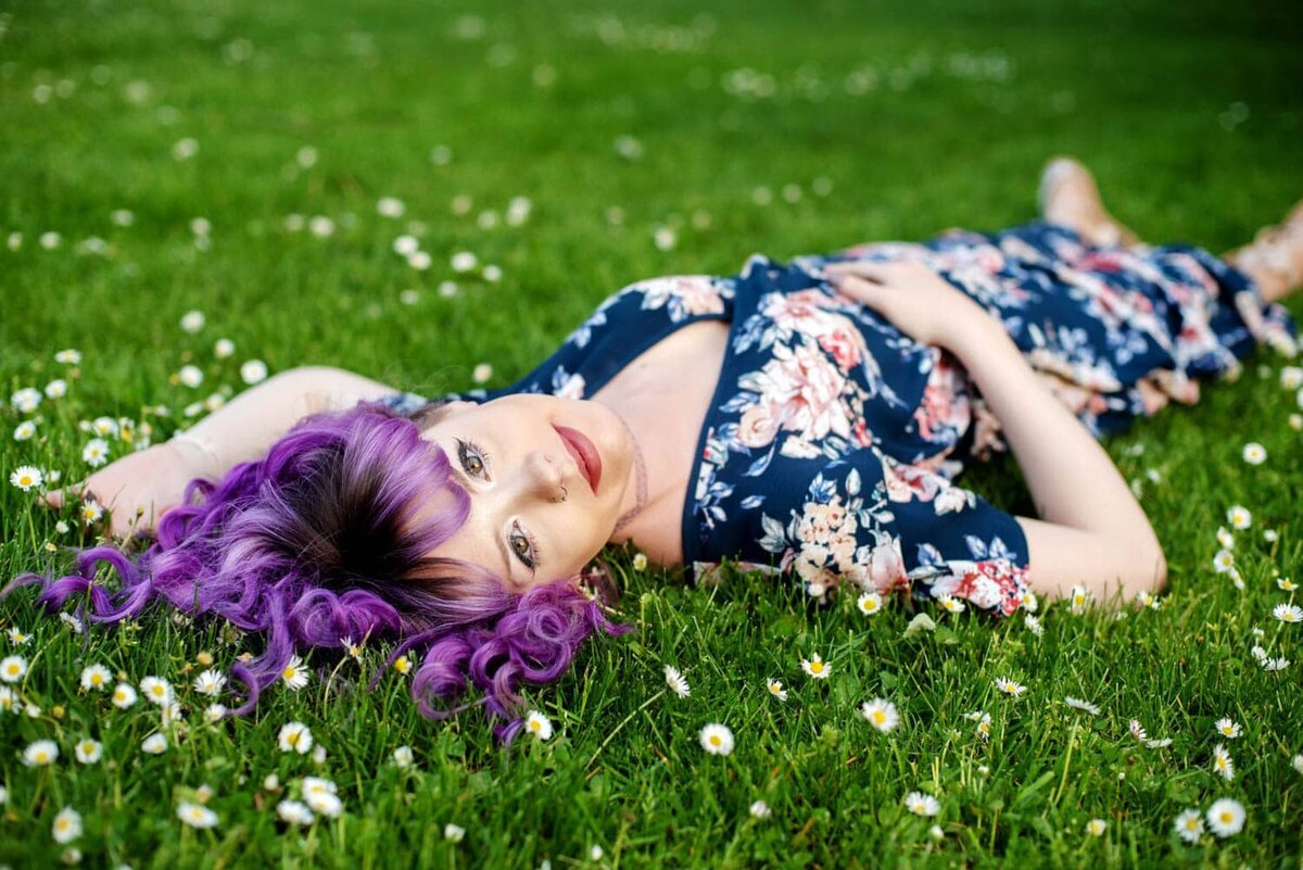 Colorful senior photo of girl with purple curly hair and a floral dress,  laying in the grass surrounded by white daisies.