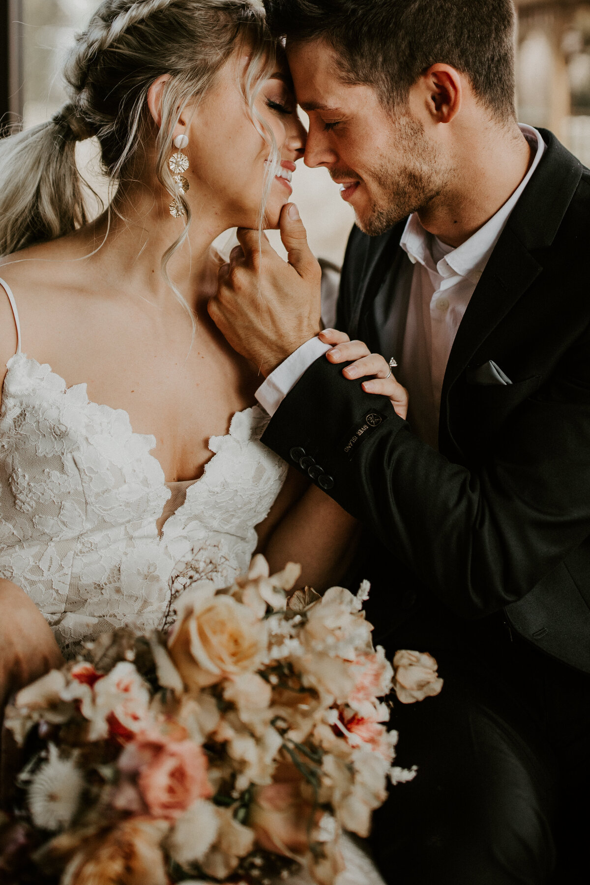 A bride and groom wearing a white wedding gown and black suit lean in for a kiss with blush and yellow bouquet of flowers.