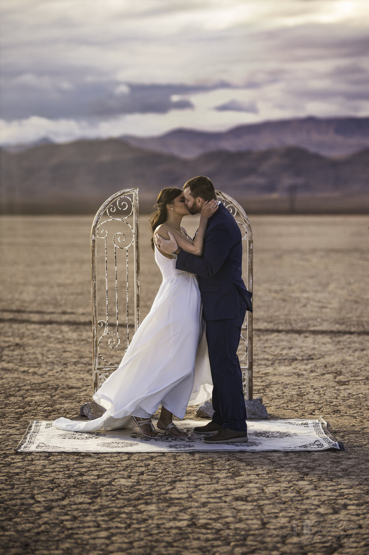 Bride and groom say I do First Kiss as man and wife purple mountains Dry lake Bed elopement Blue Suit on Groom  flowers by michelle  bride in cream color wedding dress with deep  plunging  neckline mountain skyline  sunset las vegas wedding photographers mk delacy photography