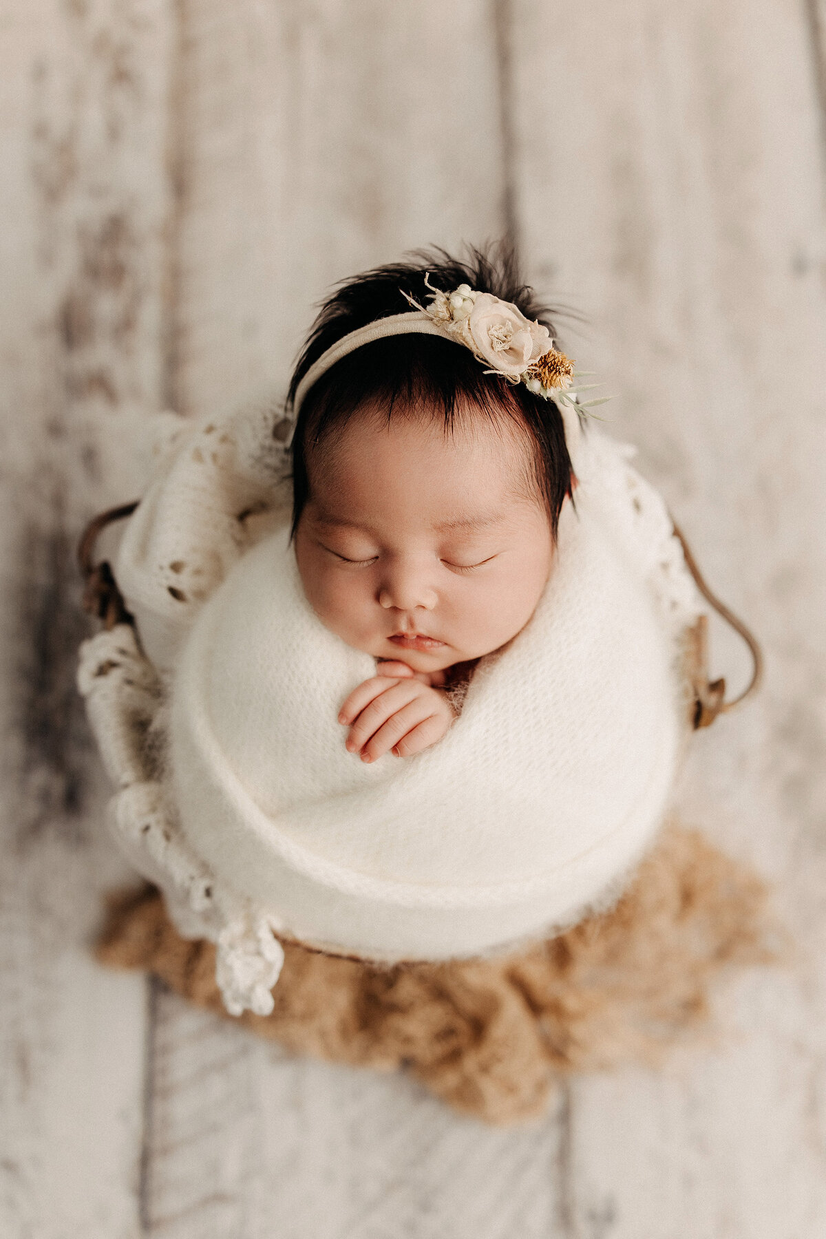 Newborn baby girl wrapped in white knit swaddle with floral headband, sleeping in basket during San Diego newborn photo session.