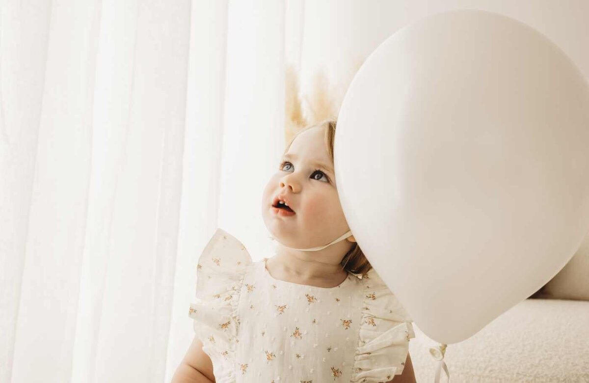 A Ruua Hamdan Photography image of a curious toddler in a floral dress looking up at a large white balloon. Soft, gentle lighting.