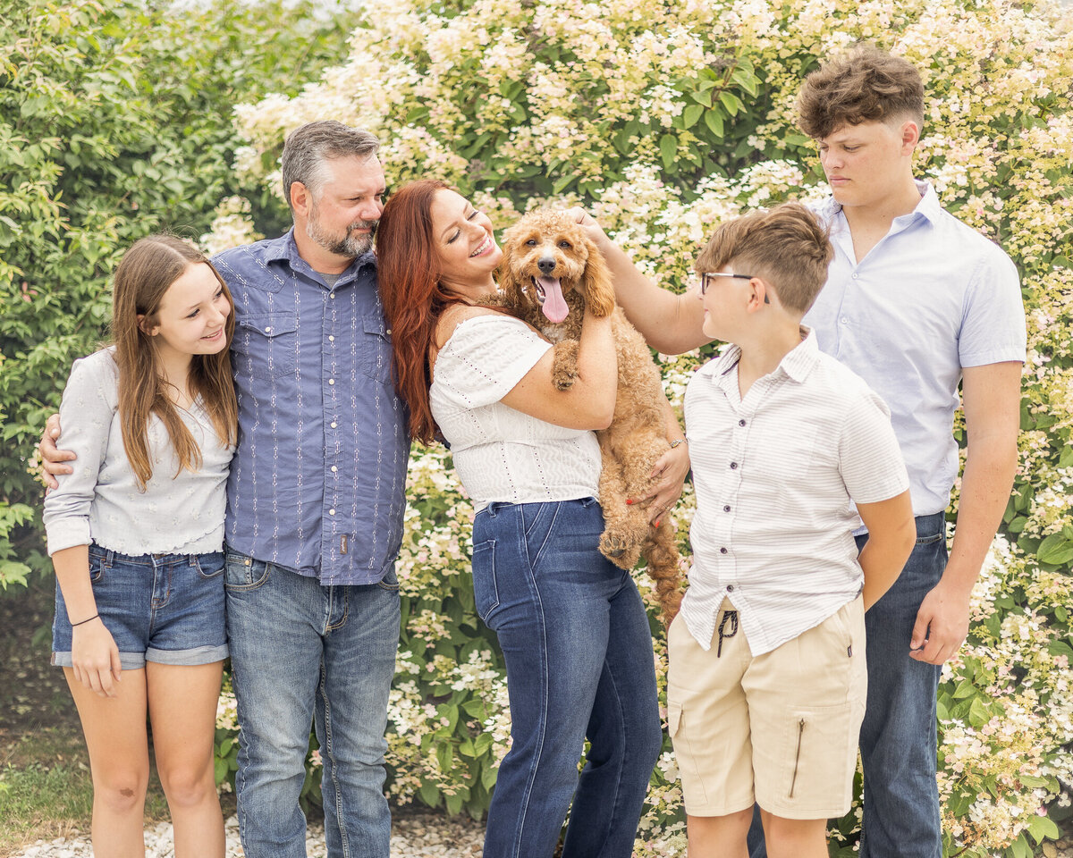 Family of five plus their light brown dog are standing in front of a bush. Everyone is looking at the dog smiling.  Daughter is wearing blue jean shorts, a long sleeve light blue shirt, white shoes, and has long light brown hair.  Dad is wearing blue jeans, blue and white stripped button up shirt, and has short pepper hair and beard.  Younger son is wearing a light tan button up and khaki shorts, white shoes, short light brown hair.  Oldest son is wearing a light blue short sleeve button up, blue jeans, and has short light brown hair