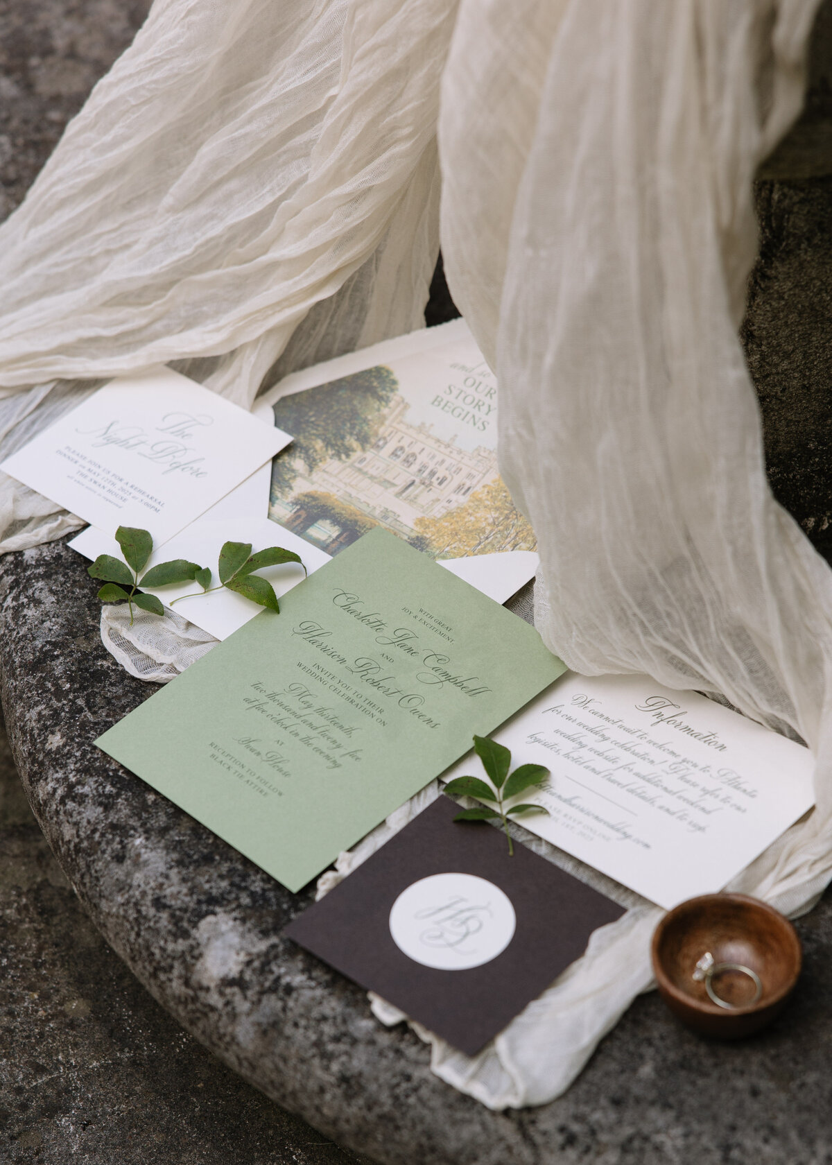 Flat lay of an elegant wedding invitation suite with green, white, and black stationery, styled with flowing ivory fabric, fresh greenery, and a wooden ring dish, photographed by Kayt Zirkle Photography.