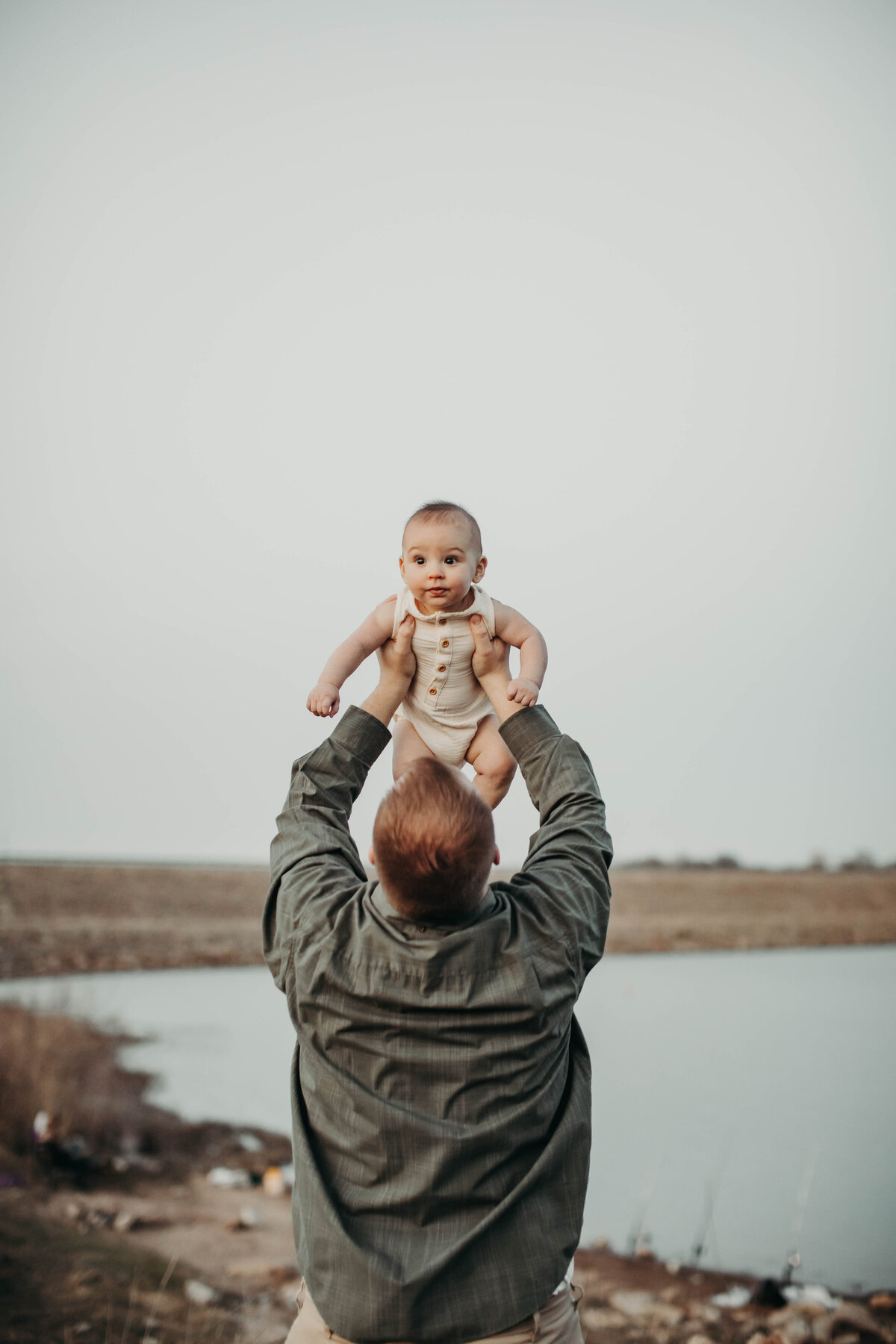 Dad holding baby in the air in front of lake. 