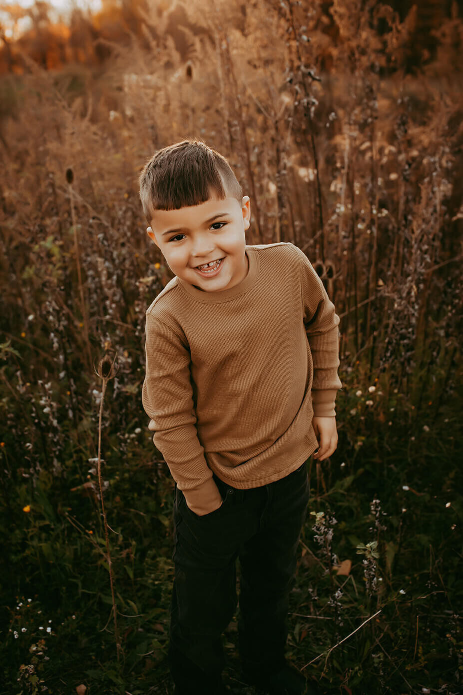 a five year old boy with his hand in his pocket looking and smiling at the camera in a field