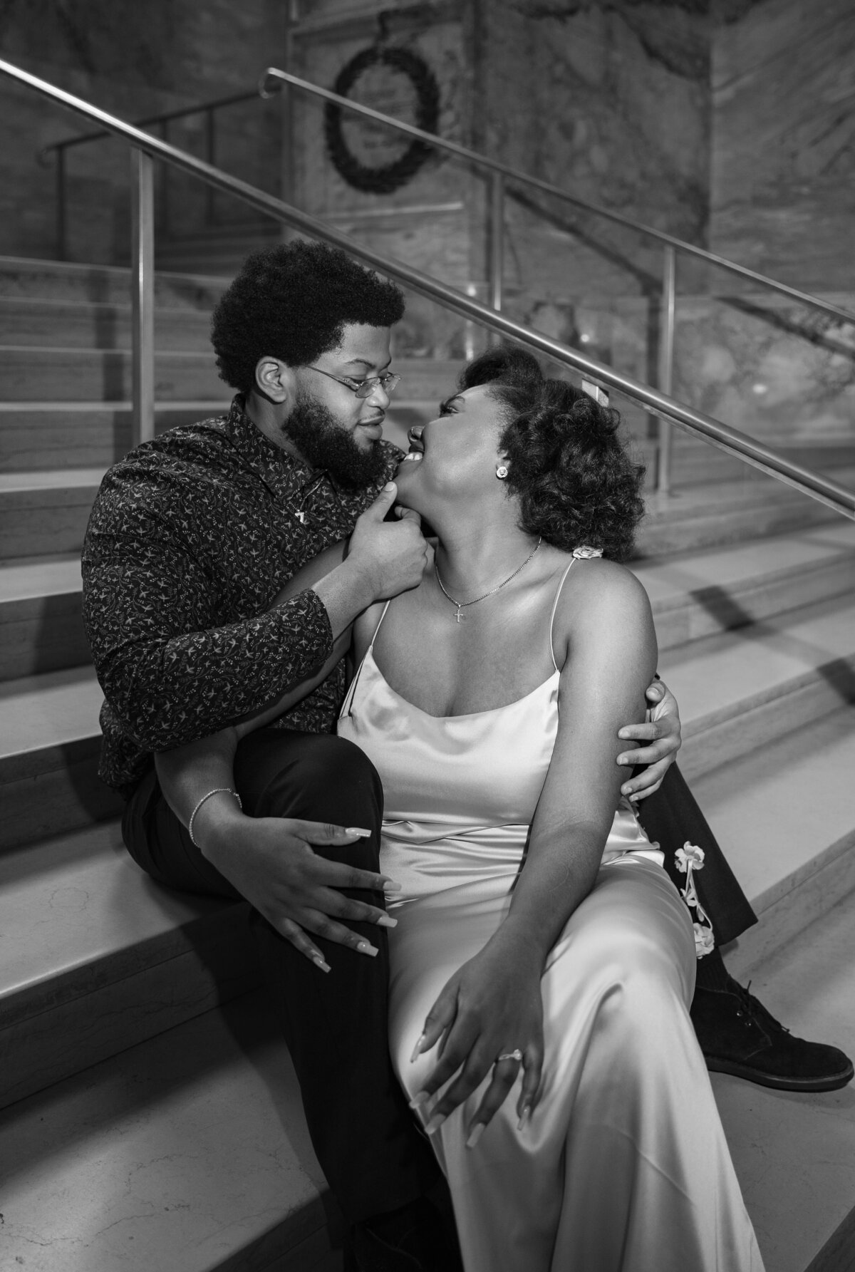 Wedding Photographers in RI | Kelsey Sheehan Photography Timeless Rhode Island Weddings | A couple sits on stairs, smiling affectionately at each other. He gently holds her chin; she wears a satin dress. The scene conveys warmth and intimacy.