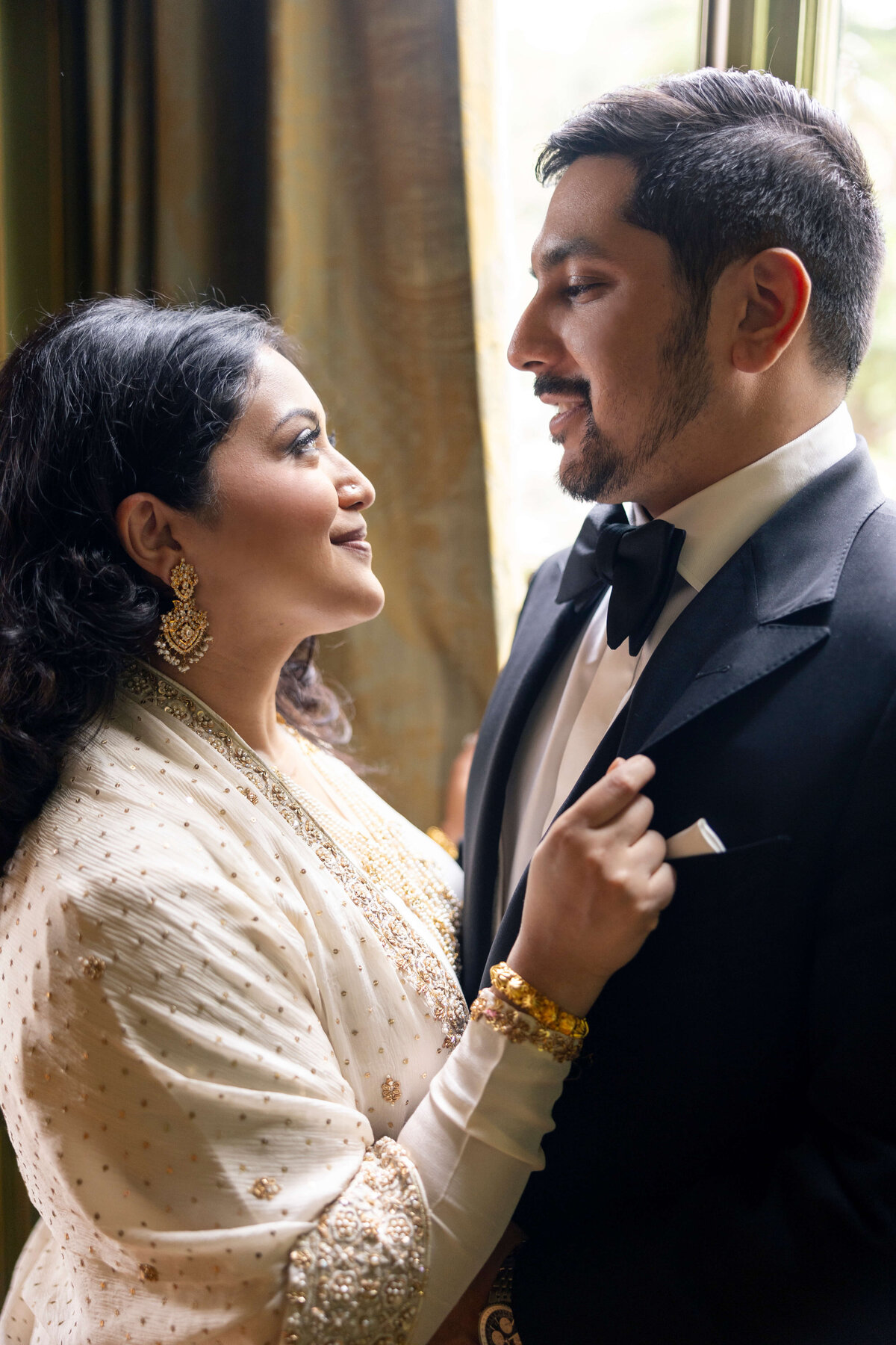 A couple stands closely, gazing into each others eyes. The woman wears an elegant white saree with gold details and earrings, while the man is in a black tuxedo with a bow tie. Soft light filters through a window behind them.