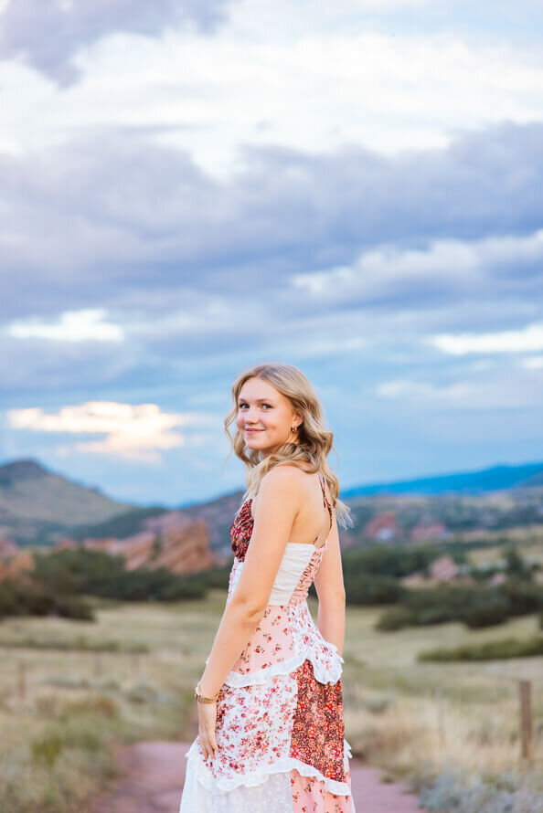 Senior girl looks over her shoulder with red rocks and blue sky behind her.