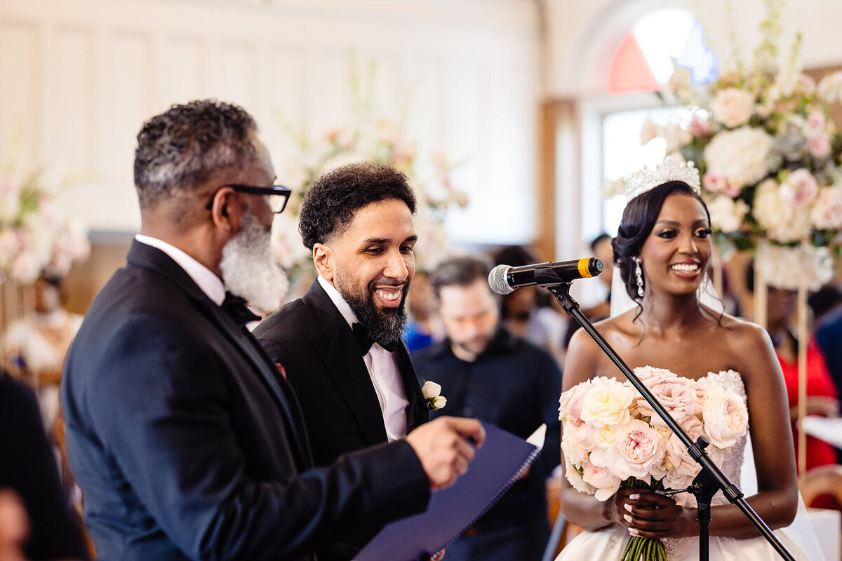 A happy moment captured as a groom smiles while speaking at a microphone beside a bride during a wedding ceremony, with guests in the background
