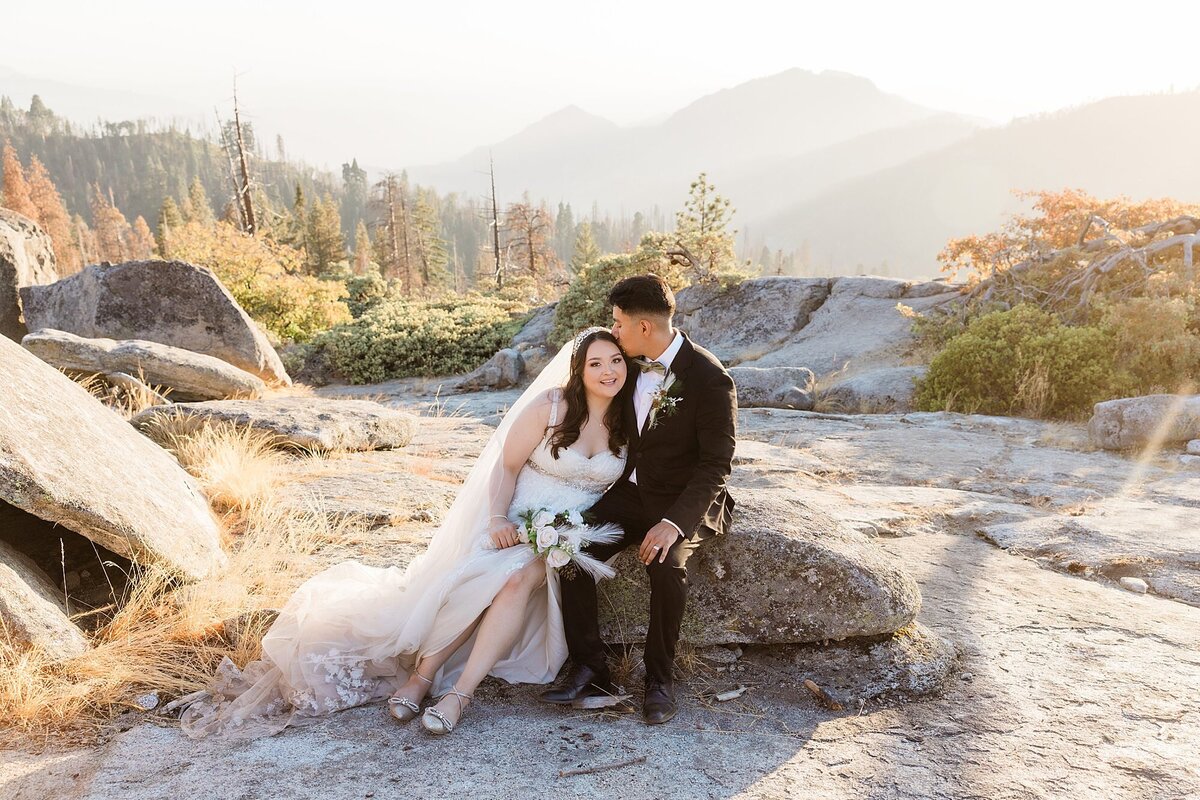 The bride and groom pose on a rock with the mountains in the background during sunset.