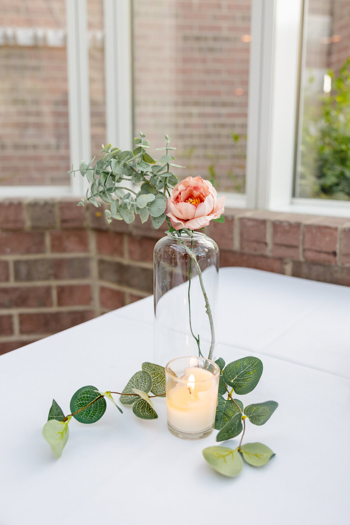 A simple and elegant reception table with a glass vase, a single pink flower, greenery, and a flickering candle.