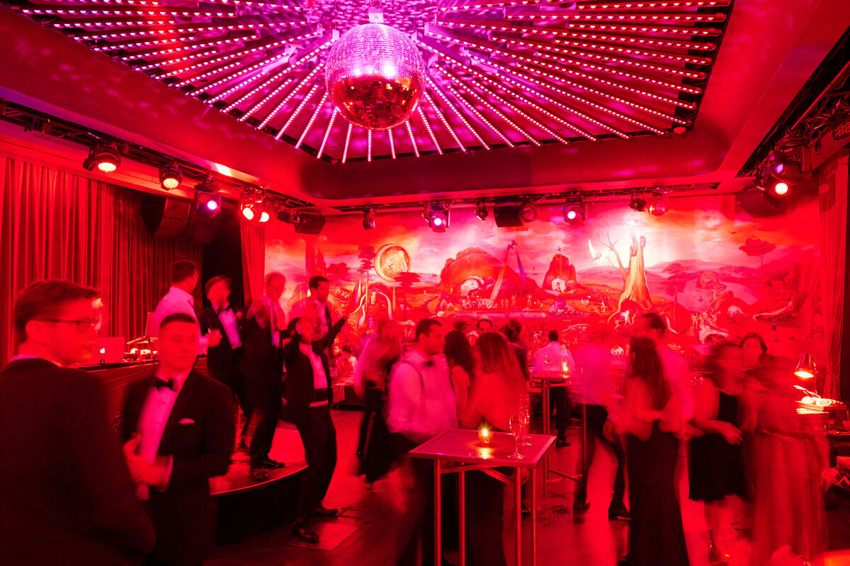 This image was captured at the New York Public Library in New York City by photographer Shawn Connell. The scene depicts a lively crowd dressed in formal attire dancing under a large, illuminated disco ball in a vibrant, red-lit room. In the background, a colorful mural with fantasy elements adds to the festive atmosphere, and several cocktail tables are scattered around the dance floor. Aaron Navak Films served as Videographers for this event.