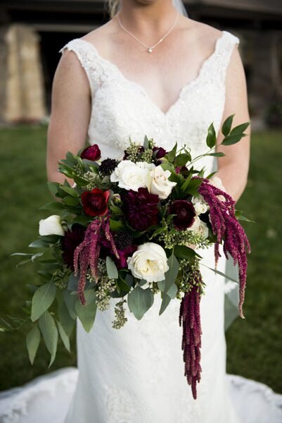 A cropped in shot of a bride holding her bouquet featuring dark burgundy florals and white roses.