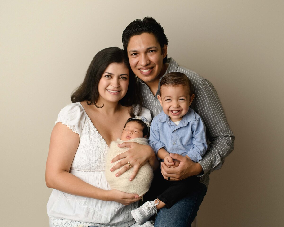Mom, Dad, and big brother pose with their newborn baby girl for her Menifee newborn photoshoot. Mom, dad, and big brother are all smiling at the camera as mom is holding the baby girl.
