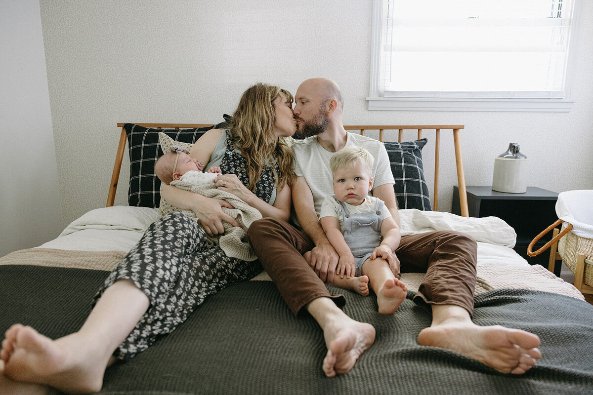 Family piles onto mom and dad's bed for candid photos while mom and dad give a kiss to each other. 