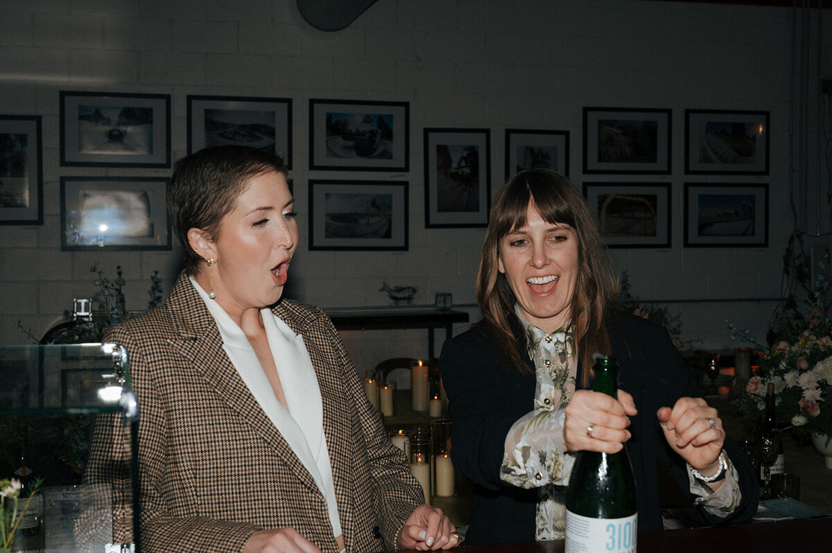 Two women smile while one of them pops open a bottle of champagne.