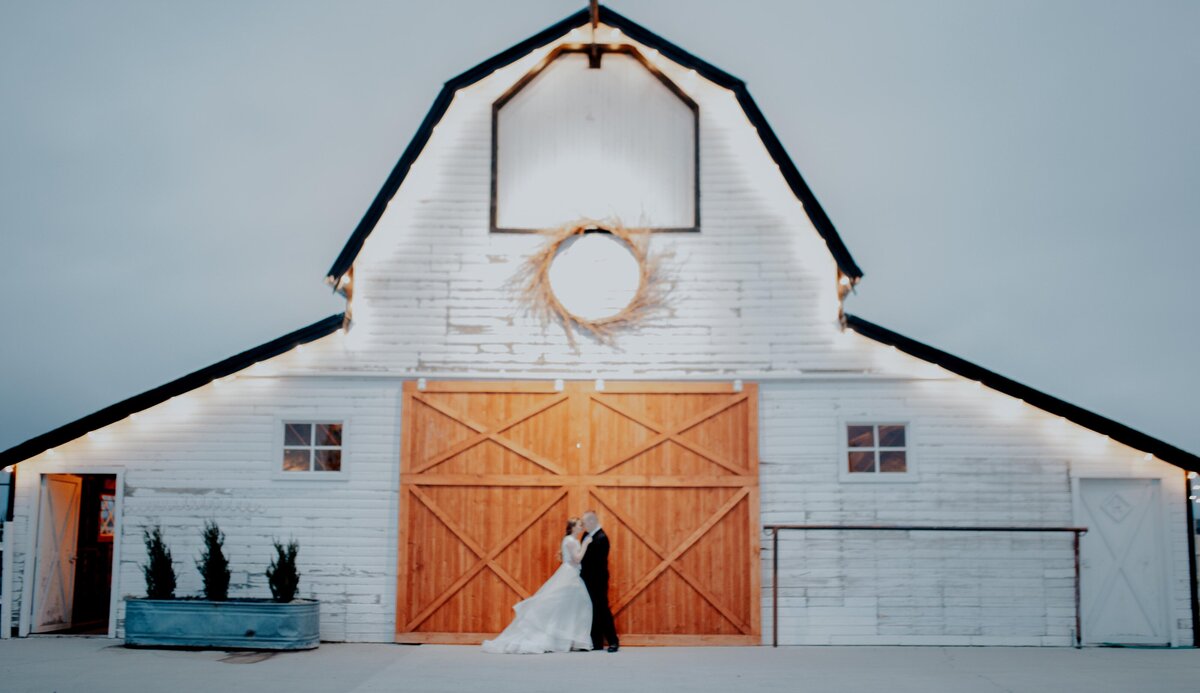 white barn wedding in Idaho Falls with bride and groom embracing each other outside of the barn doors with the barn lit up as the sun is set