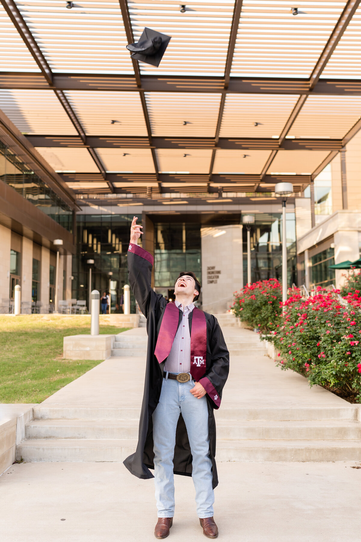 Texas A&M senior guy throwing up grad cap in the air while wearing gown and Aggie stole in front of Zachry Engineering Complex