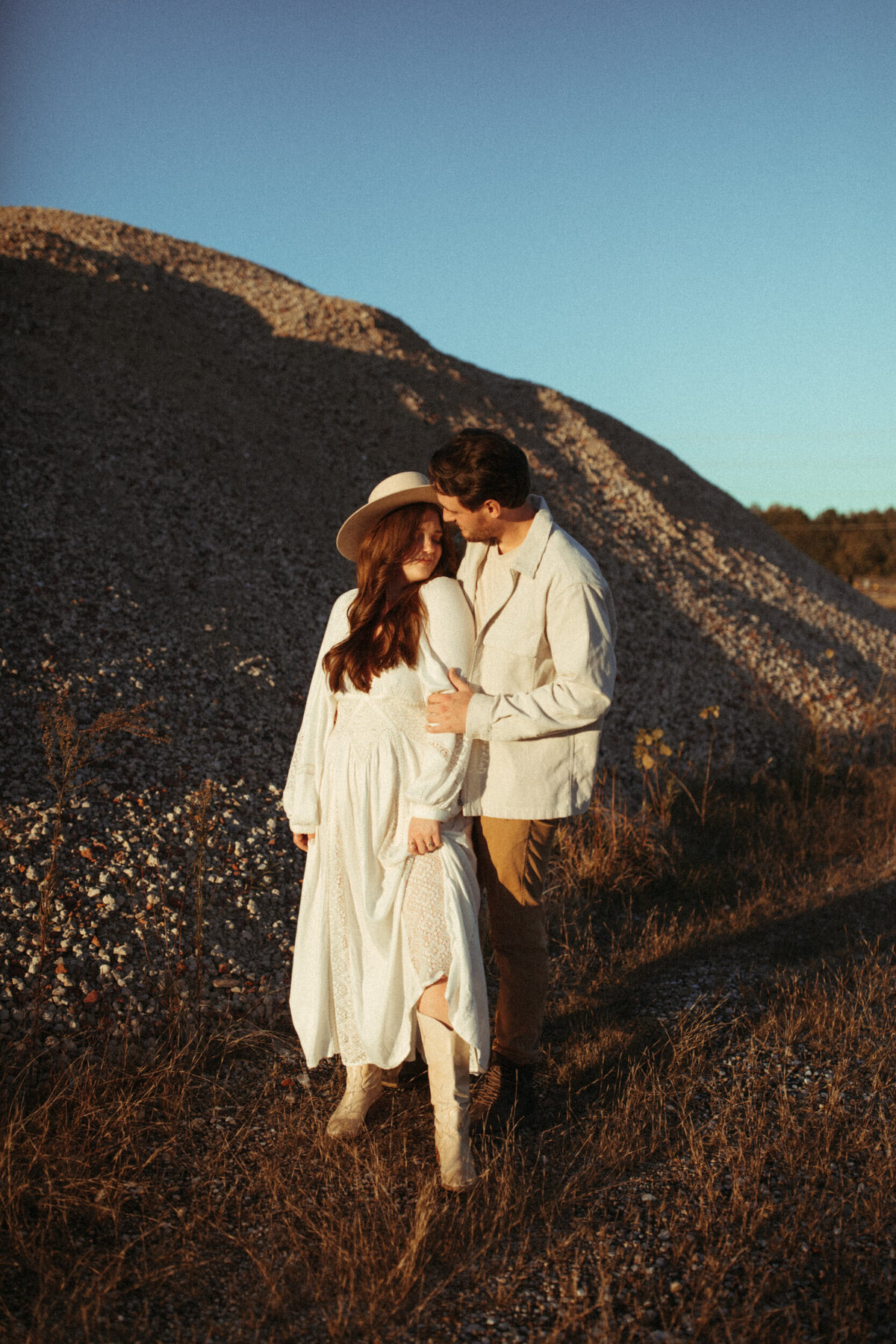 Girl in long white dress, tall white boots and a boho hat standing next to her husband in a white jacket and tan pants in a gravel pit