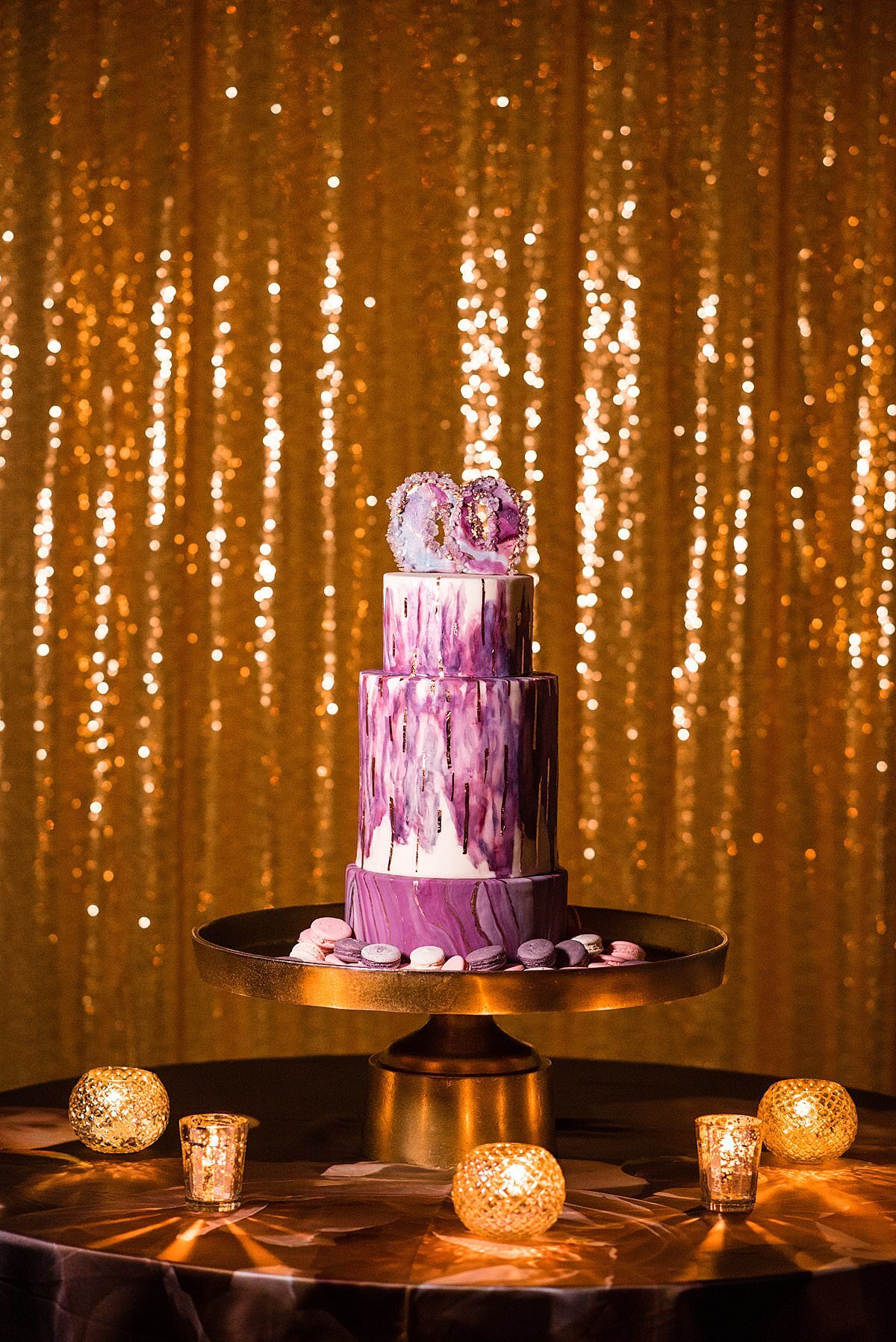 purple art decor wedding cake with geode topper surrounded by pink and purple macrons with a gold sequin drapery backdrop