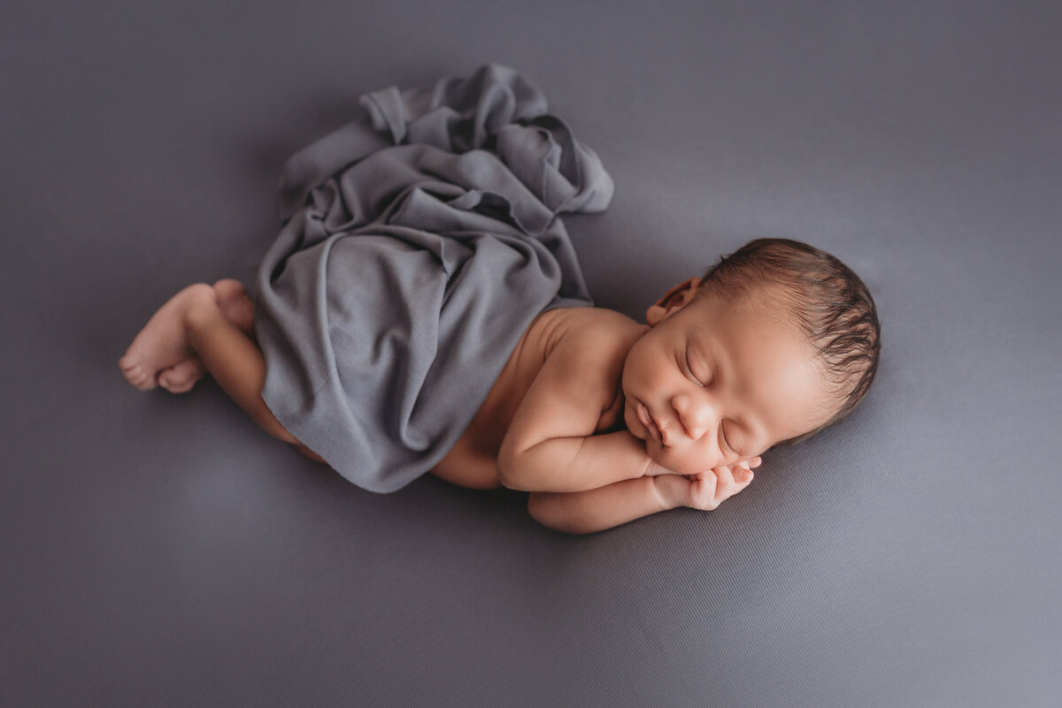 Newborn baby boy asleep and posed on blue fabric on side with hands under his cheek, feet together and blue wrap covering his diaper