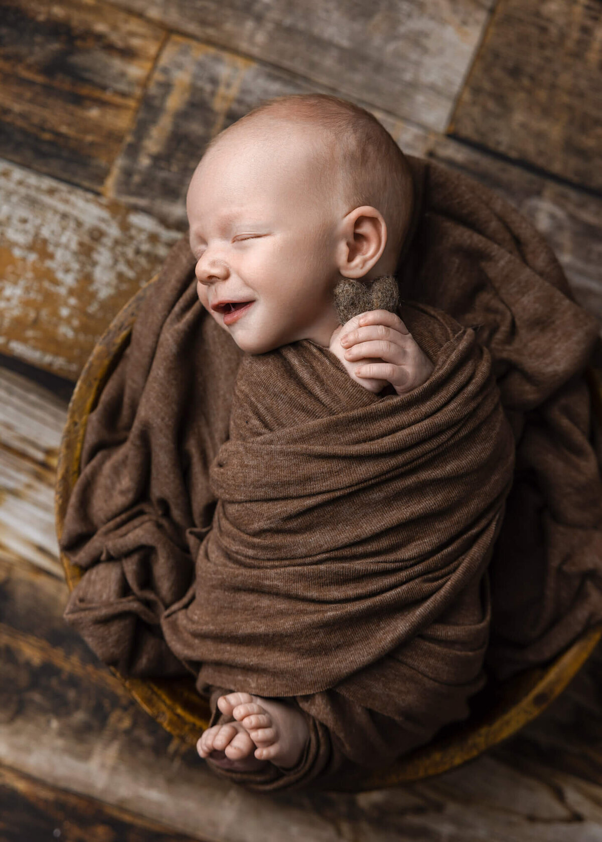 smiling newborn baby wrapped in brown fabric  asleep in a wooden bowl  holding a felted brown heart